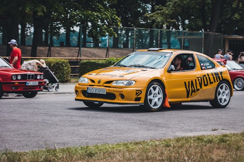 A vibrant yellow rally car with 'Valvoline' branding is moving along a road. It has a prominent front bumper and a visible rear spoiler. There's a male driver inside wearing a helmet. Behind the rally car, a red classic car is parked, with a man wearing a hat pushing a stroller to the left. The background consists of lush green trees and a park setting with a fenced area.