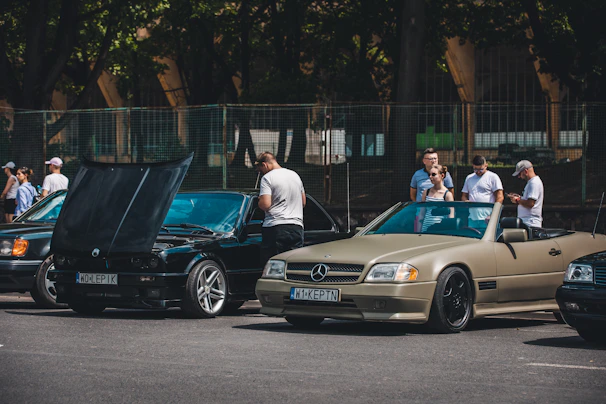 A group of diverse drivers chatting and smiling in front of their cars in a sunny parking lot.
