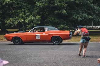 A person with a red backpack is seen taking a photograph of an orange vintage car driving on a road. The background consists of lush green trees, which set a natural backdrop for the scene.