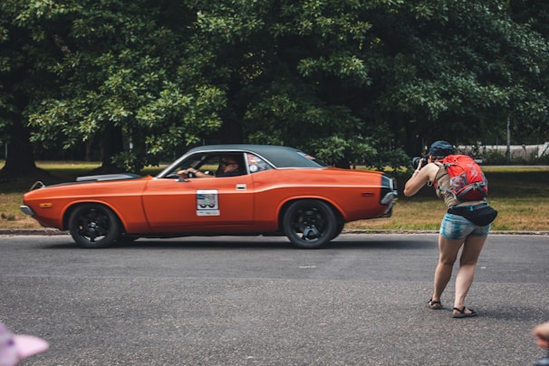 A person with a red backpack is seen taking a photograph of an orange vintage car driving on a road. The background consists of lush green trees, which set a natural backdrop for the scene.
