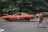 A person with a red backpack is seen taking a photograph of an orange vintage car driving on a road. The background consists of lush green trees, which set a natural backdrop for the scene.
