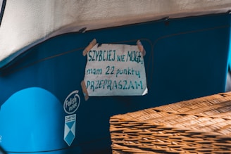 A handwritten sign is taped to the side of a blue vehicle or object. The sign is made of paper with text in Polish, secured with pieces of tape at the corners. There is a wicker basket or similar object partially visible in the foreground.