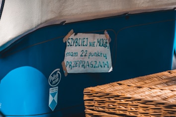 A handwritten sign is taped to the side of a blue vehicle or object. The sign is made of paper with text in Polish, secured with pieces of tape at the corners. There is a wicker basket or similar object partially visible in the foreground.