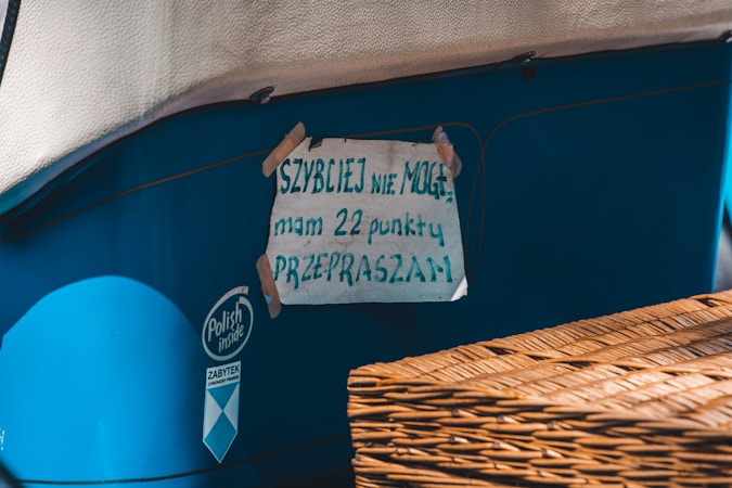 A handwritten sign is taped to the side of a blue vehicle or object. The sign is made of paper with text in Polish, secured with pieces of tape at the corners. There is a wicker basket or similar object partially visible in the foreground.