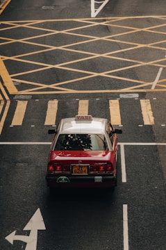 A red taxi car is seen from above at an intersection with clearly marked yellow grid lines and road markings. The car's roof is white, and it is driving on a black asphalt road.