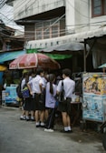 A group of students in uniforms stand closely together near a street vendor under large umbrellas. The scene is set against an urban backdrop with visible power lines and a building.