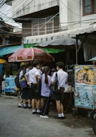 A group of students in uniforms stand closely together near a street vendor under large umbrellas. The scene is set against an urban backdrop with visible power lines and a building.