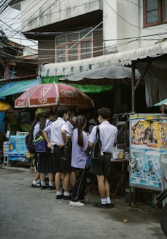 A group of students in uniforms stand closely together near a street vendor under large umbrellas. The scene is set against an urban backdrop with visible power lines and a building.