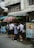 A group of students in uniforms stand closely together near a street vendor under large umbrellas. The scene is set against an urban backdrop with visible power lines and a building.
