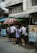 A group of students in uniforms stand closely together near a street vendor under large umbrellas. The scene is set against an urban backdrop with visible power lines and a building.