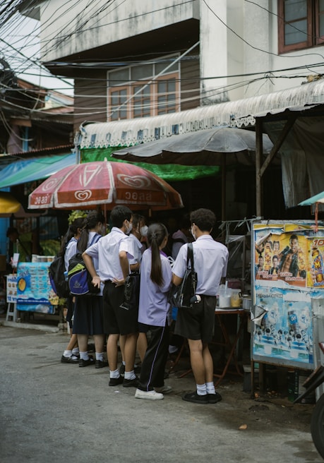 A group of students in uniforms stand closely together near a street vendor under large umbrellas. The scene is set against an urban backdrop with visible power lines and a building.