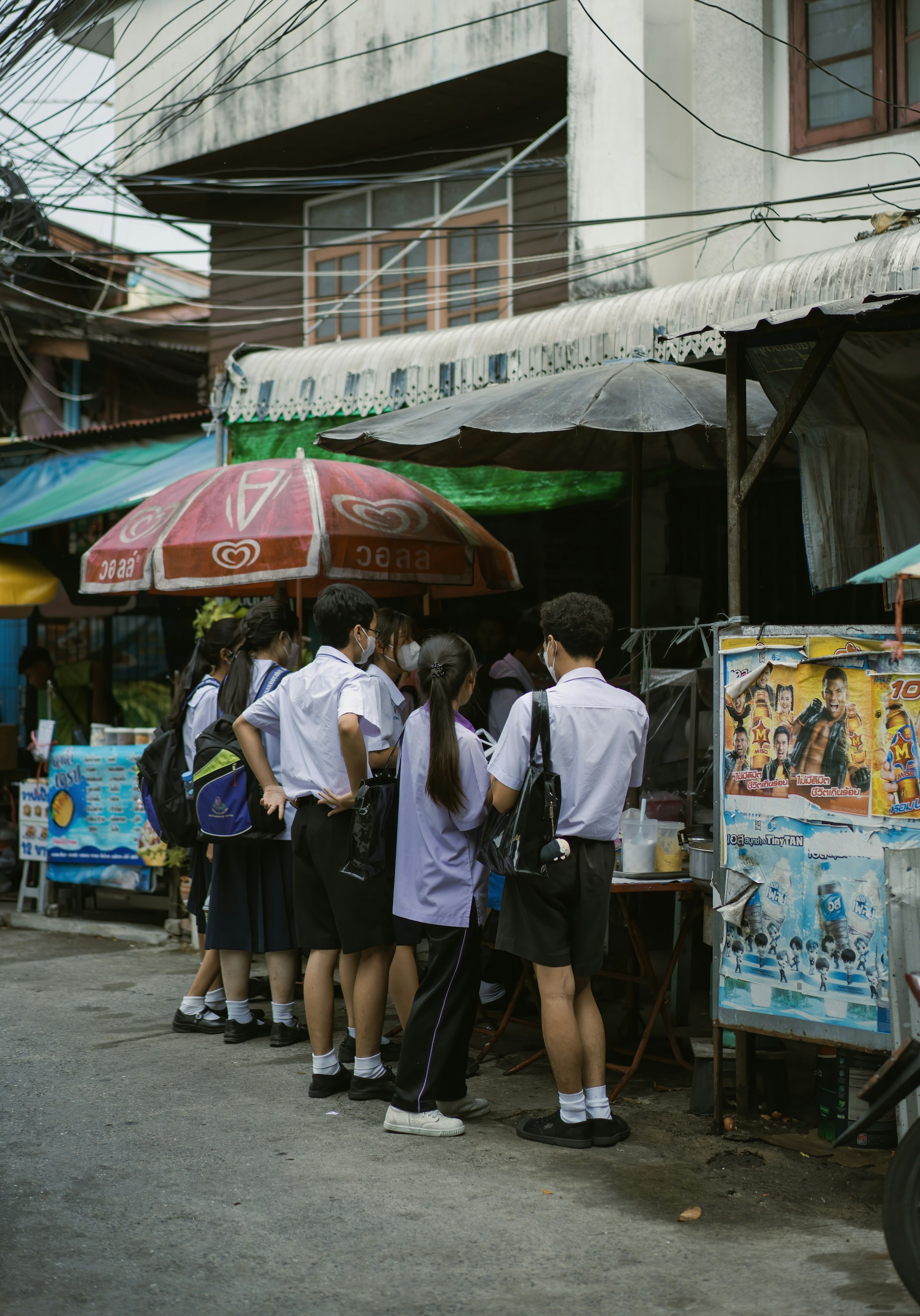 A group of students in uniforms stand closely together near a street vendor under large umbrellas. The scene is set against an urban backdrop with visible power lines and a building.