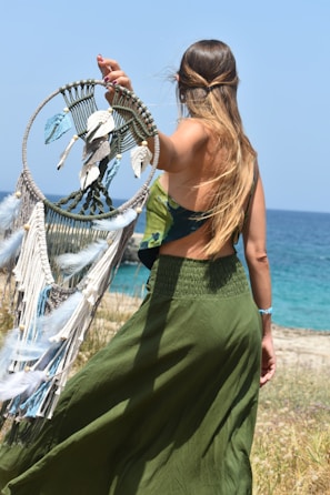 A woman stands with her back to the camera, holding a dreamcatcher adorned with feathers and beads. She is near a seaside, wearing a green flowing skirt and a sleeveless top. Her hair is long and loose, with a bohemian style.