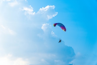 A person is paragliding against a backdrop of a clear blue sky with some fluffy white clouds. The paraglider is colorful and the scene is expansive, giving a sense of freedom and adventure.