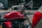 A red Royal Enfield motorcycle is parked alongside cars on a street. The image captures the details of the motorcycle's tank and handlebar, highlighting its classic design with chrome elements. The background is softly blurred, giving prominence to the motorcycle.