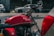 A red Royal Enfield motorcycle is parked alongside cars on a street. The image captures the details of the motorcycle's tank and handlebar, highlighting its classic design with chrome elements. The background is softly blurred, giving prominence to the motorcycle.