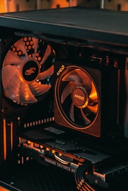 A close-up view of a computer case interior featuring a cooling fan with the brand 'Deep Cool' and a CPU cooler marked 'AMD'. Below the components is a graphics card labeled 'Inno3D GeForce RTX'. The lighting in the image has warm orange tones, highlighting the sleek and high-tech look of the hardware.