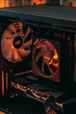 A close-up view of a computer case interior featuring a cooling fan with the brand 'Deep Cool' and a CPU cooler marked 'AMD'. Below the components is a graphics card labeled 'Inno3D GeForce RTX'. The lighting in the image has warm orange tones, highlighting the sleek and high-tech look of the hardware.