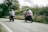 Happy couple riding motorbikes along a coastal road lined with lush greenery.