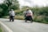 Happy couple riding motorbikes along a coastal road lined with lush greenery.
