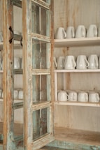 A rustic wooden cabinet with a distressed finish, featuring glass panels and iron handles. Inside, two shelves are neatly lined with white ceramic mugs.
