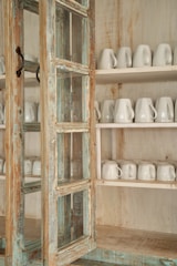 A rustic wooden cabinet with a distressed finish, featuring glass panels and iron handles. Inside, two shelves are neatly lined with white ceramic mugs.