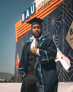 A person wearing a graduation cap and gown is standing confidently in front of a modern building with geometric patterns and a visible sign reading 'BAU'. The background also displays flags and mountains in the distance.