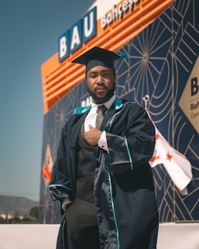 A person wearing a graduation cap and gown is standing confidently in front of a modern building with geometric patterns and a visible sign reading 'BAU'. The background also displays flags and mountains in the distance.