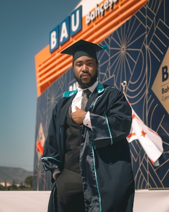 A person wearing a graduation cap and gown is standing confidently in front of a modern building with geometric patterns and a visible sign reading 'BAU'. The background also displays flags and mountains in the distance.