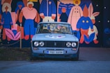An older model car with a faded paint job is parked on a street. Behind the car, there is a large, colorful mural depicting various stylized human figures and a rabbit. The mural is composed of vibrant colors including pink, blue, and orange. The license plate of the car reads '790 AKM'.