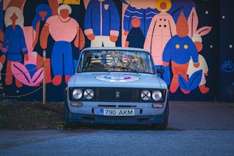 An older model car with a faded paint job is parked on a street. Behind the car, there is a large, colorful mural depicting various stylized human figures and a rabbit. The mural is composed of vibrant colors including pink, blue, and orange. The license plate of the car reads '790 AKM'.