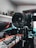 A camera mounted on a tripod is prominently placed in a room with a computer setup in the background. The camera lens is facing towards the viewer, with reflections visible on the lens glass. The room is equipped with a monitor, keyboard, and various tech accessories on a desk. There are plants and figurines on the shelf in the background.