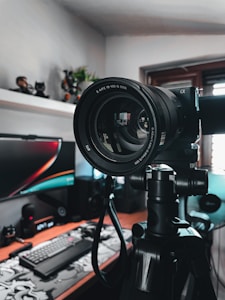 A camera mounted on a tripod is prominently placed in a room with a computer setup in the background. The camera lens is facing towards the viewer, with reflections visible on the lens glass. The room is equipped with a monitor, keyboard, and various tech accessories on a desk. There are plants and figurines on the shelf in the background.