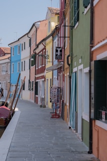 A picturesque street lined with colorful houses in shades of blue, red, yellow, green, and orange. The buildings have windows with shutters, and a small canal runs alongside with boats moored. There is a display stand and trolley on the sidewalk, and a sign in Italian hangs from the building.