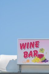 A bright and colorful sign displaying the words 'Wine Bar' with artistic illustrations of a wine glass and grapes. The sign is mounted on a structure against a clear blue sky backdrop.