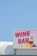 A bright and colorful sign displaying the words 'Wine Bar' with artistic illustrations of a wine glass and grapes. The sign is mounted on a structure against a clear blue sky backdrop.