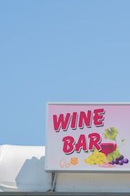 A bright and colorful sign displaying the words 'Wine Bar' with artistic illustrations of a wine glass and grapes. The sign is mounted on a structure against a clear blue sky backdrop.