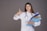 A woman in a white lab coat with a stethoscope around her neck is holding a stack of colorful folders in one hand and giving a thumbs-up with the other. She has long brown hair and is smiling confidently against a plain gray background.