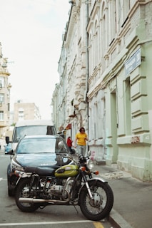 A vintage motorcycle is parked on a narrow city street beside a dark-colored car. A person in a yellow shirt walks along the sidewalk next to old buildings with ornate architecture. The street is lined with multi-story buildings, and a sign with Cyrillic script is attached to one.