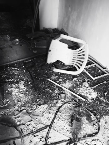 A broken plastic chair is lying on its side amidst scattered debris on the floor. The ground is covered with dust, rubble, and various broken items, including a crumpled plastic bottle and cables. The scene suggests a state of disrepair or destruction.