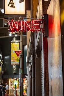 A neon sign displaying the word 'WINE' is affixed to a building on a bustling city street. The surroundings include various illuminated signs and a busy shopfront, with people walking along the sidewalk. The scene is lit with ambient urban lighting.