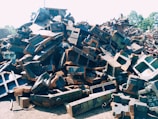A large pile of metal scrap and debris is stacked outdoors. The metal pieces are of various shapes and sizes, with some showing signs of rust and wear. Sunlight is cast over the pile, highlighting the angular shapes and textures.