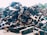 A large pile of metal scrap and debris is stacked outdoors. The metal pieces are of various shapes and sizes, with some showing signs of rust and wear. Sunlight is cast over the pile, highlighting the angular shapes and textures.