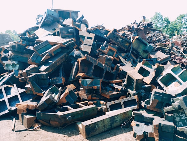 A large pile of metal scrap and debris is stacked outdoors. The metal pieces are of various shapes and sizes, with some showing signs of rust and wear. Sunlight is cast over the pile, highlighting the angular shapes and textures.