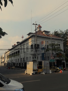 A street scene featuring a multi-story building with white walls and red-tiled roofs. The architecture shows a colonial style, and there are multiple signs and utility poles with wires in front of the building. The street is quiet, with a few construction barriers and cones visible. Trees and a parked vehicle can also be seen.