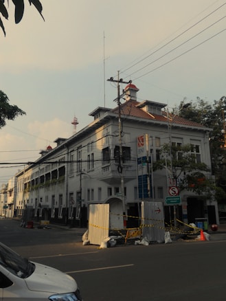 A street scene featuring a multi-story building with white walls and red-tiled roofs. The architecture shows a colonial style, and there are multiple signs and utility poles with wires in front of the building. The street is quiet, with a few construction barriers and cones visible. Trees and a parked vehicle can also be seen.