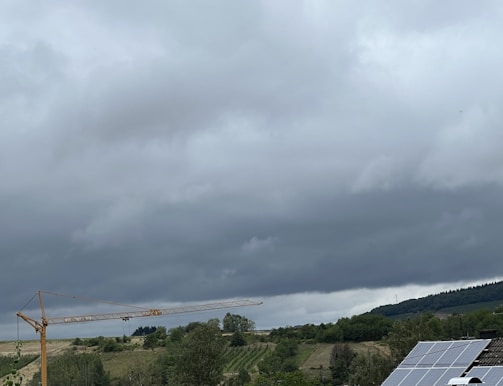 A landscape with overcast skies and a large construction crane stretching horizontally. The scene includes rolling hills with patches of green vegetation, and in the foreground, there are solar panels on a rooftop.