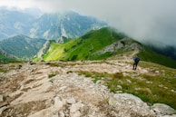A lush green mountainous landscape with a rocky trail winding through the hills. Clouds partially obscure the distant peaks, creating a misty atmosphere. A hiker with a backpack and walking stick can be seen on the path, adding a sense of exploration and adventure.