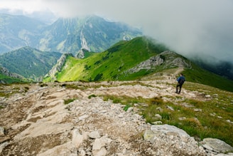 A lush green mountainous landscape with a rocky trail winding through the hills. Clouds partially obscure the distant peaks, creating a misty atmosphere. A hiker with a backpack and walking stick can be seen on the path, adding a sense of exploration and adventure.