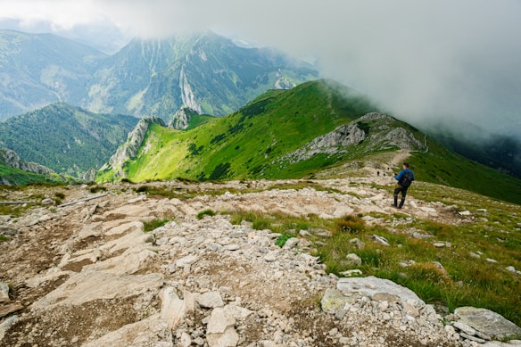 A lush green mountainous landscape with a rocky trail winding through the hills. Clouds partially obscure the distant peaks, creating a misty atmosphere. A hiker with a backpack and walking stick can be seen on the path, adding a sense of exploration and adventure.